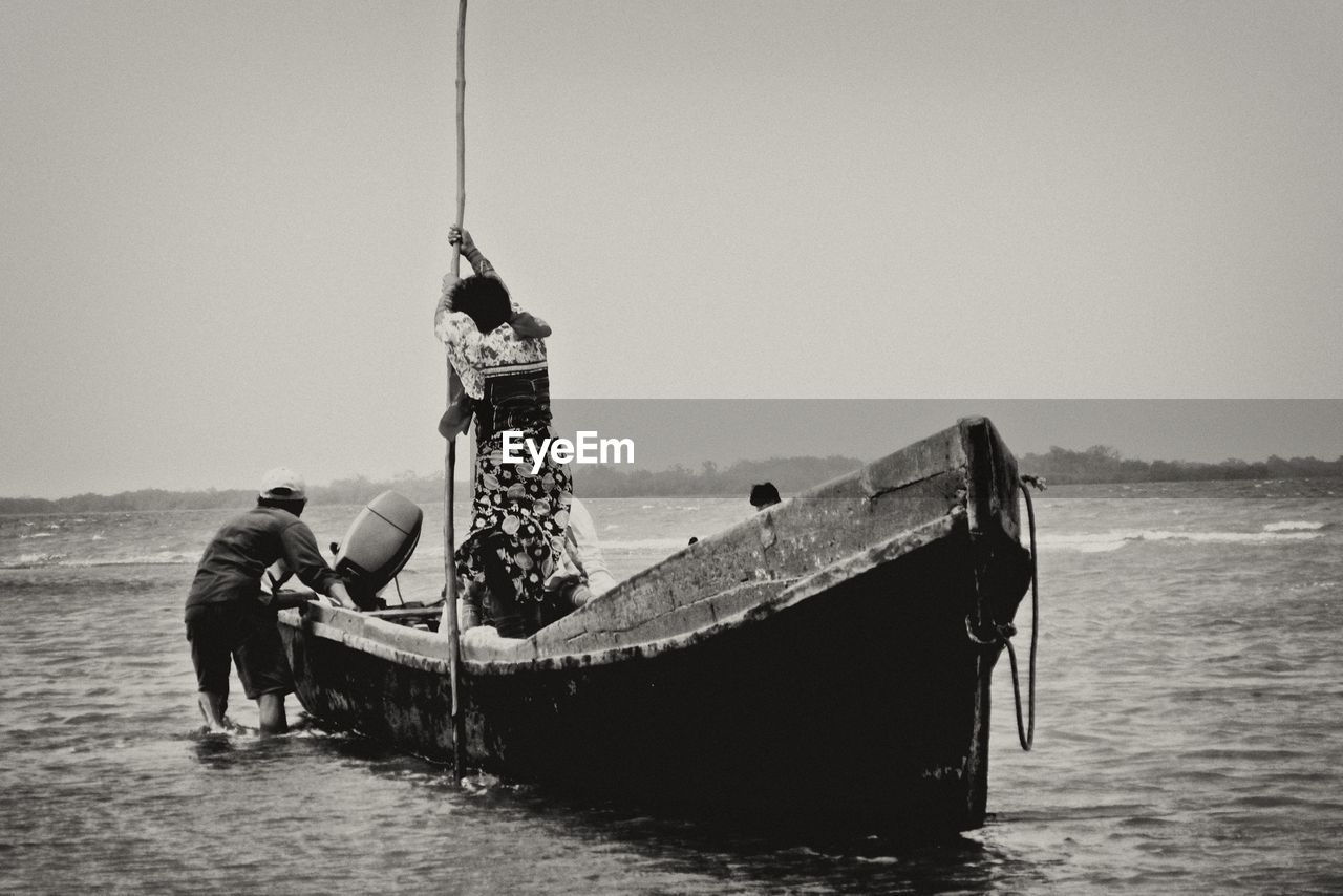 Woman holding bamboo while man pushing motorboat on sea against clear sky