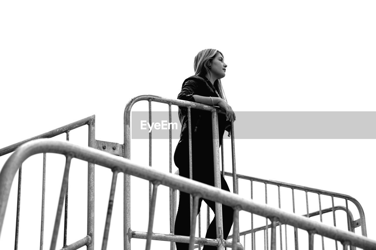 LOW ANGLE VIEW OF WOMAN STANDING BY RAILING AGAINST CLEAR SKY