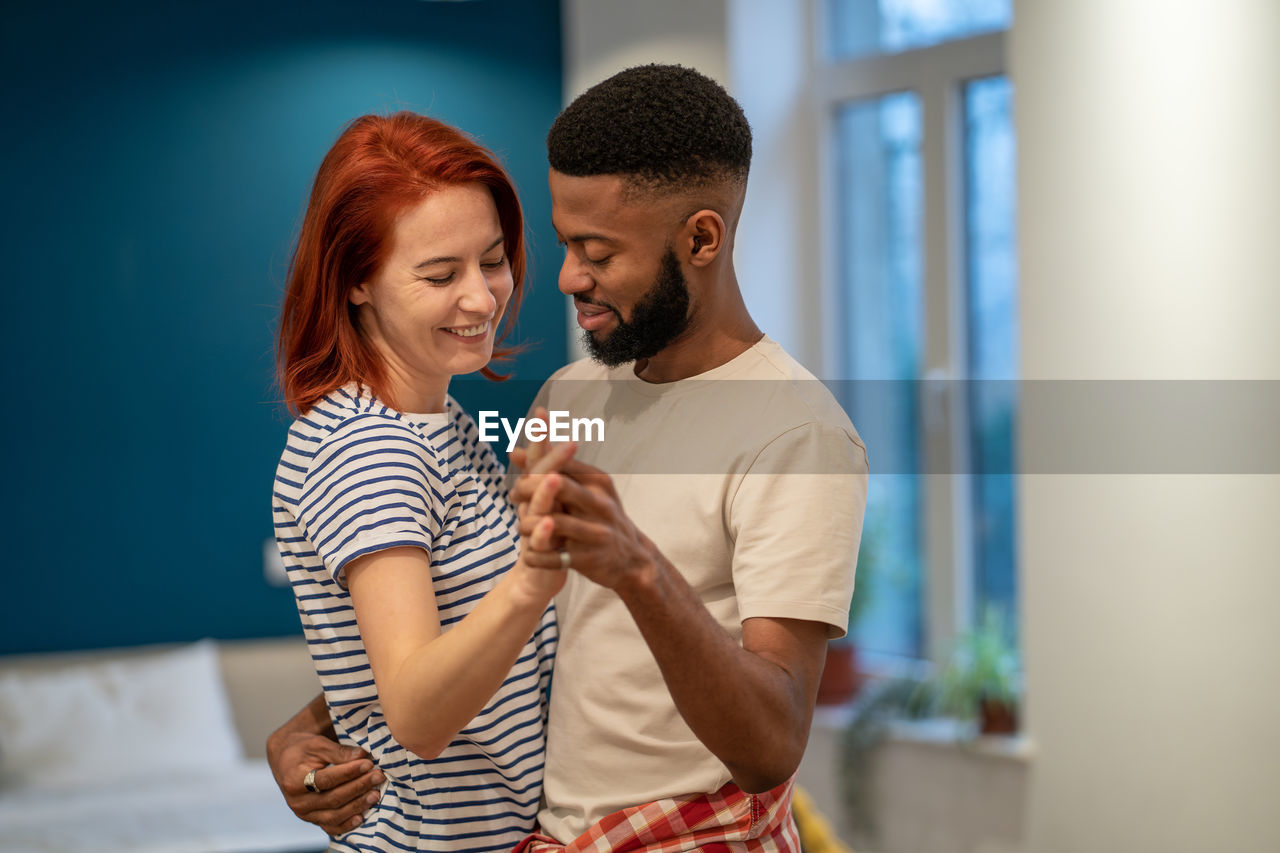 Loving young diverse couple dancing romantic dance in modern bedroom with panoramic windows.