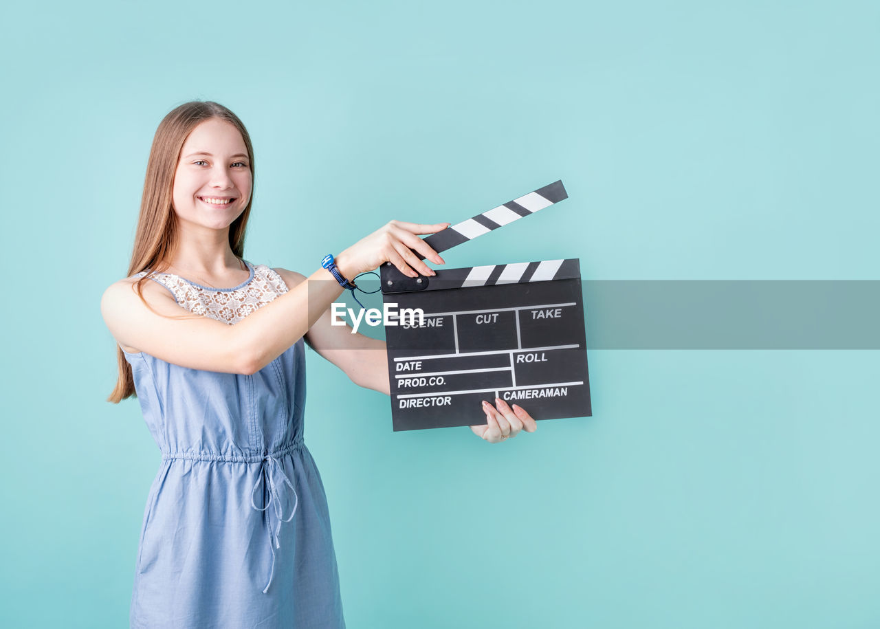 Happy smiling teenager girl in a blue dress holding a clapper board isolated on blue background 