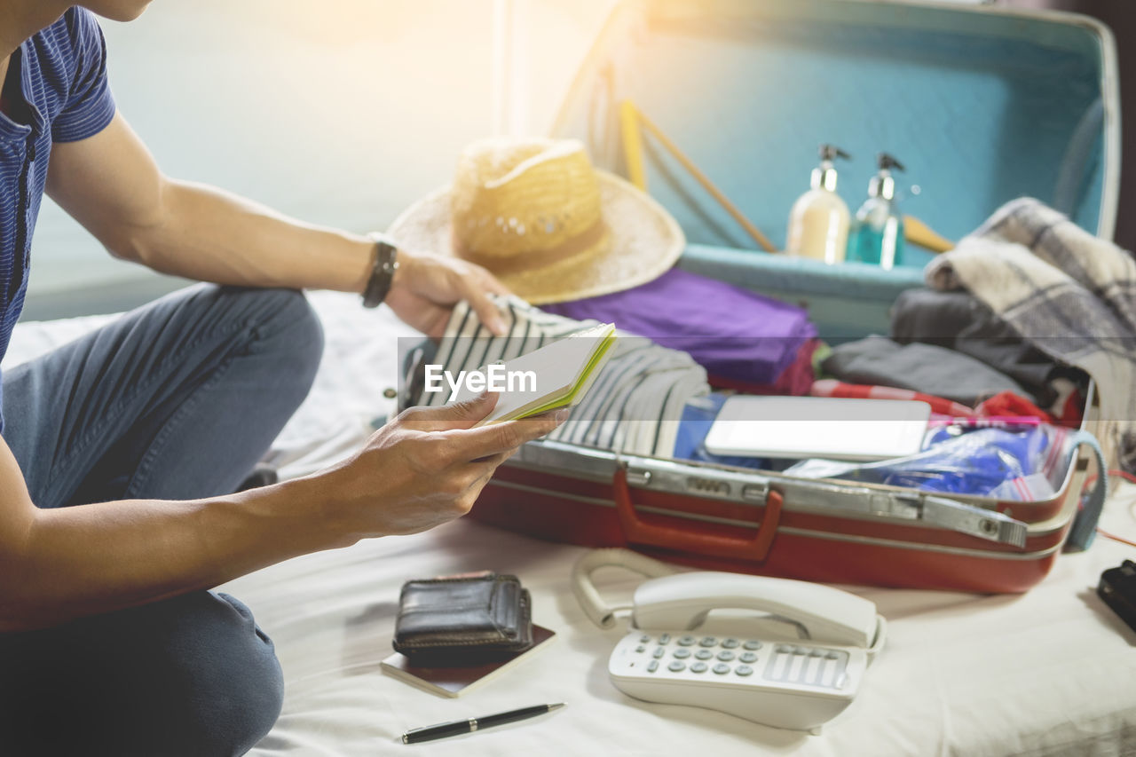 Midsection of man checking to-do-list while packing bag on bed at home