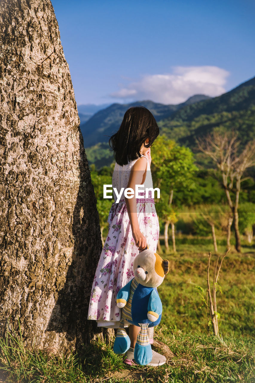 Girl holding toy while standing on grass by tree trunk against mountain and sky