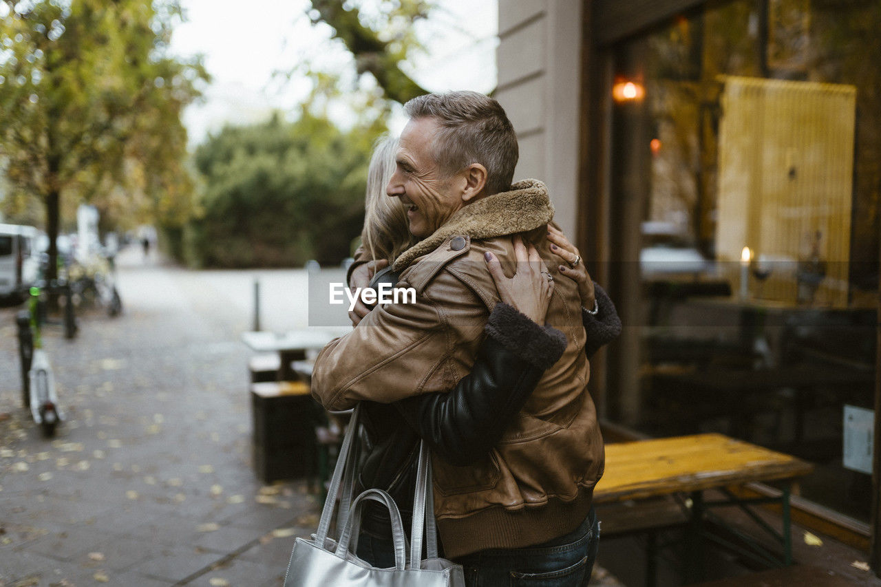 Happy man embracing woman while standing sidewalk near restaurant