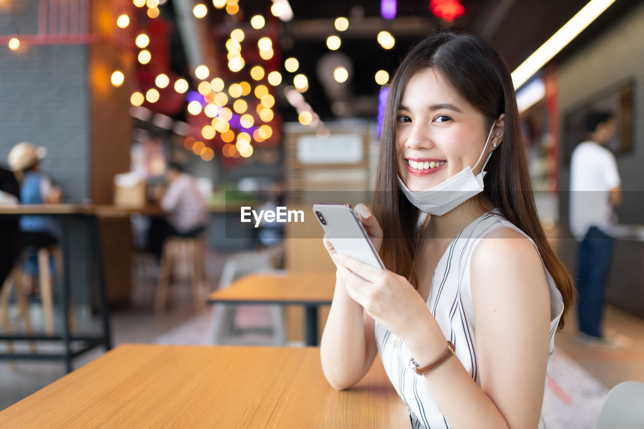 PORTRAIT OF SMILING YOUNG WOMAN HOLDING SMART PHONE WHILE USING MOBILE