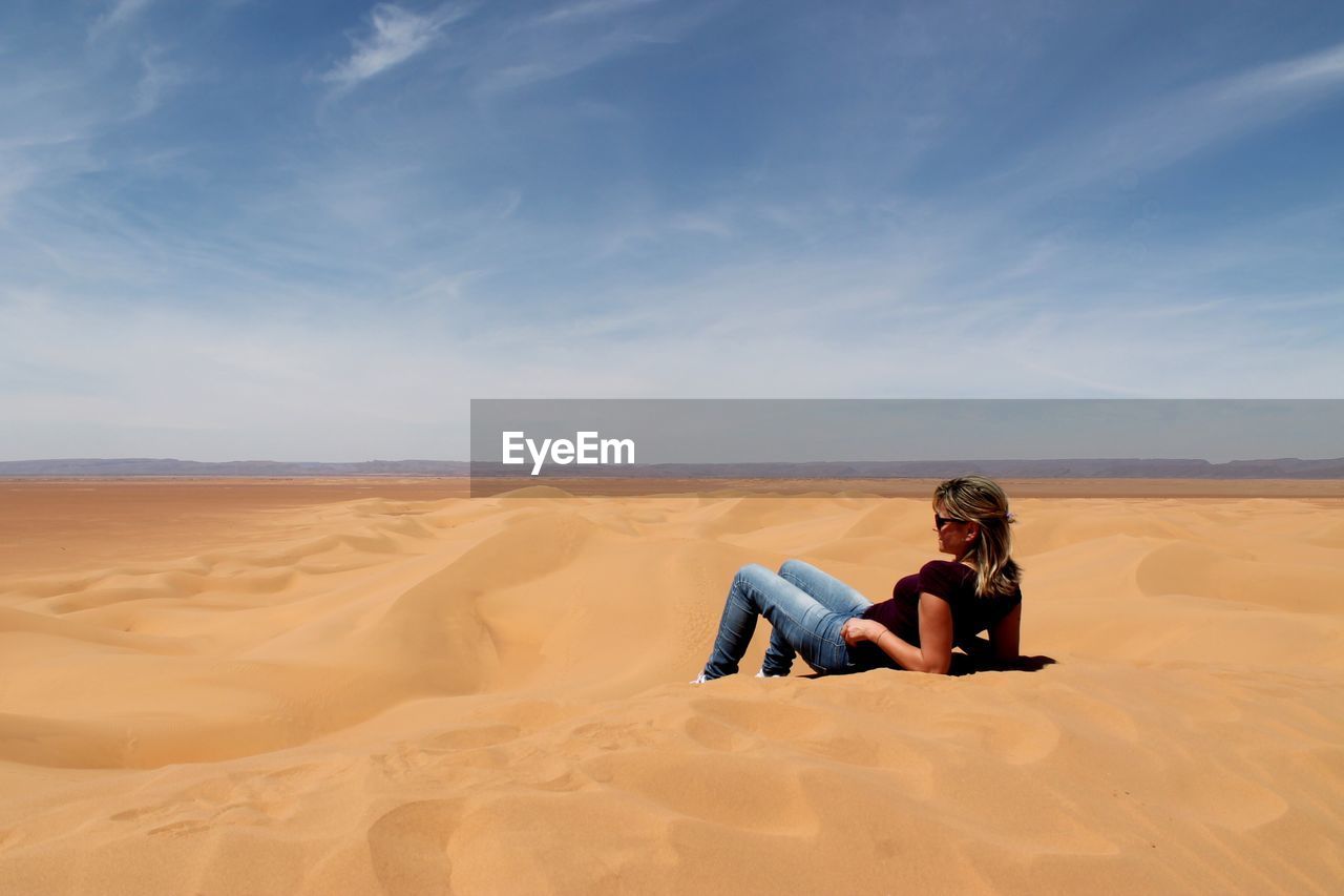 Woman relaxing on sand at desert against sky