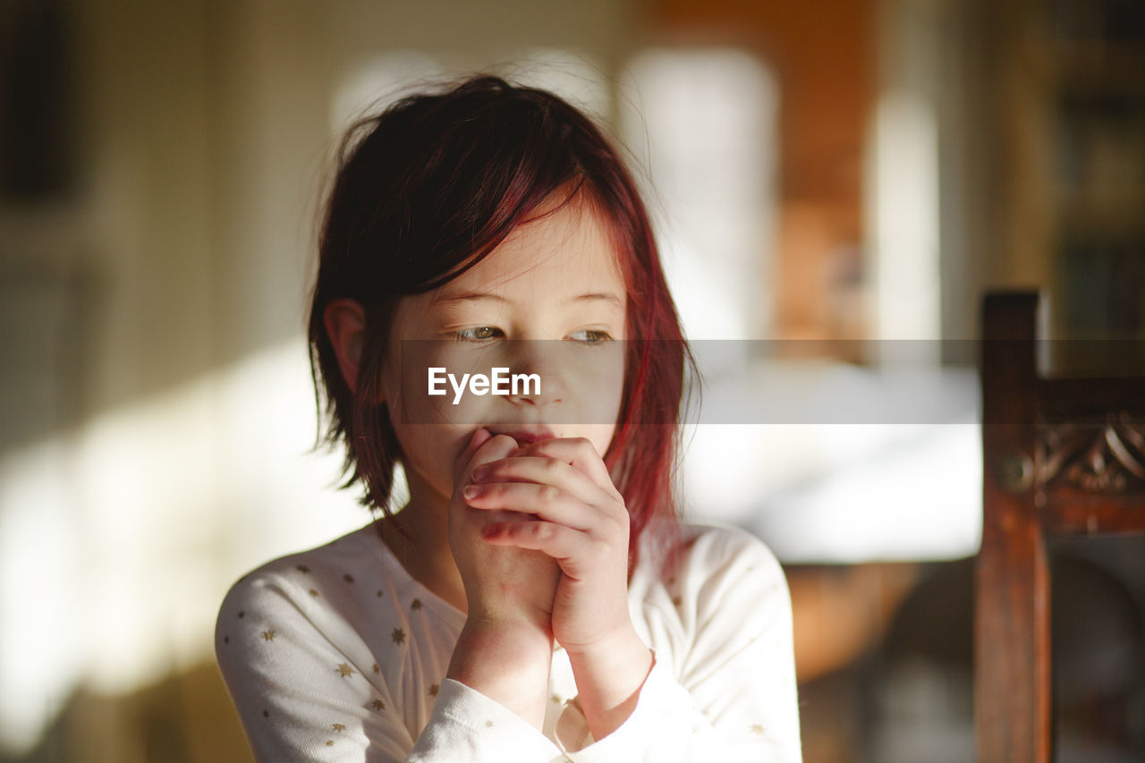 A shy little girl with dyed red hair sits alone with hands clasped