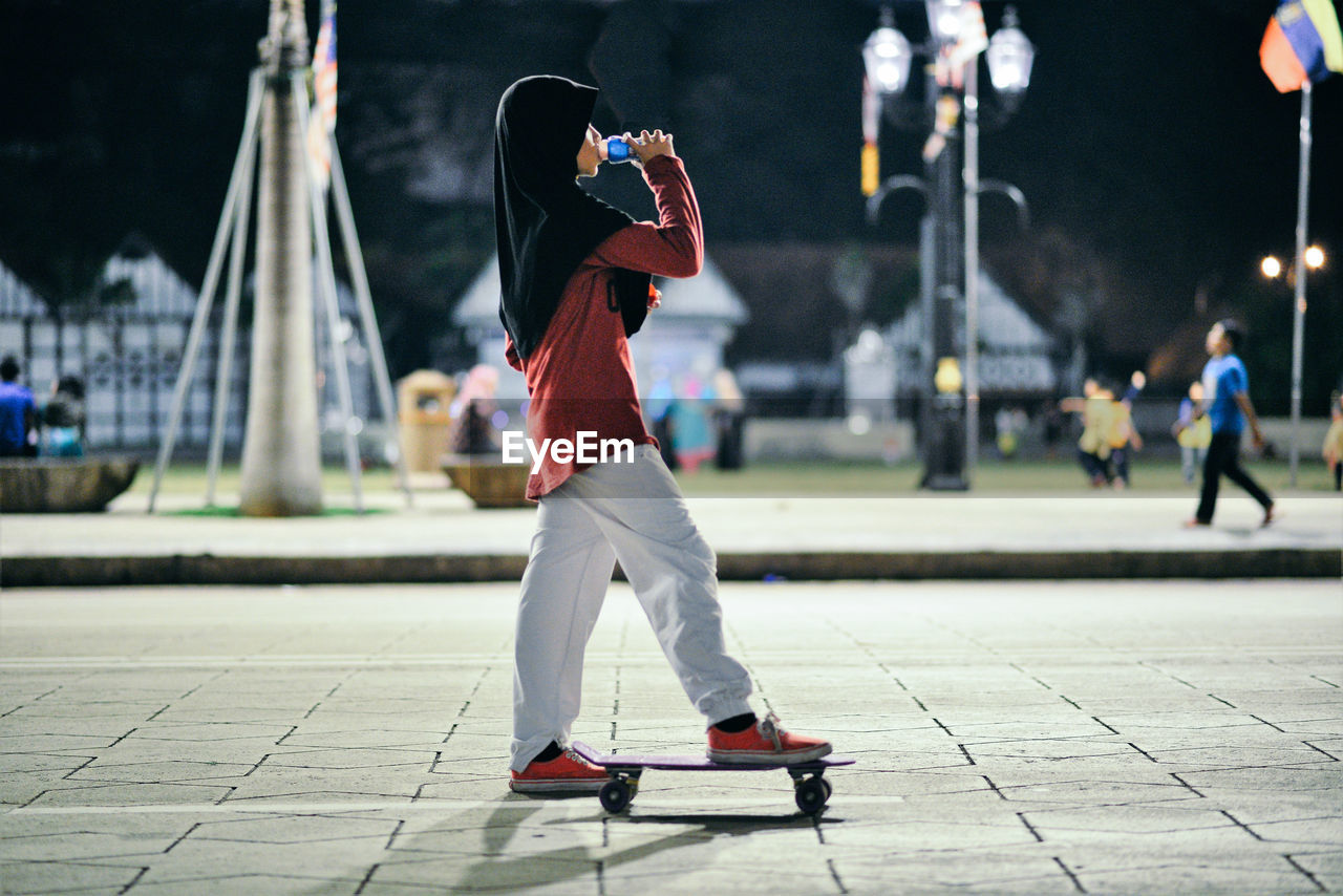 Side view of teenage girl drinking while standing with skateboard on footpath in park at night