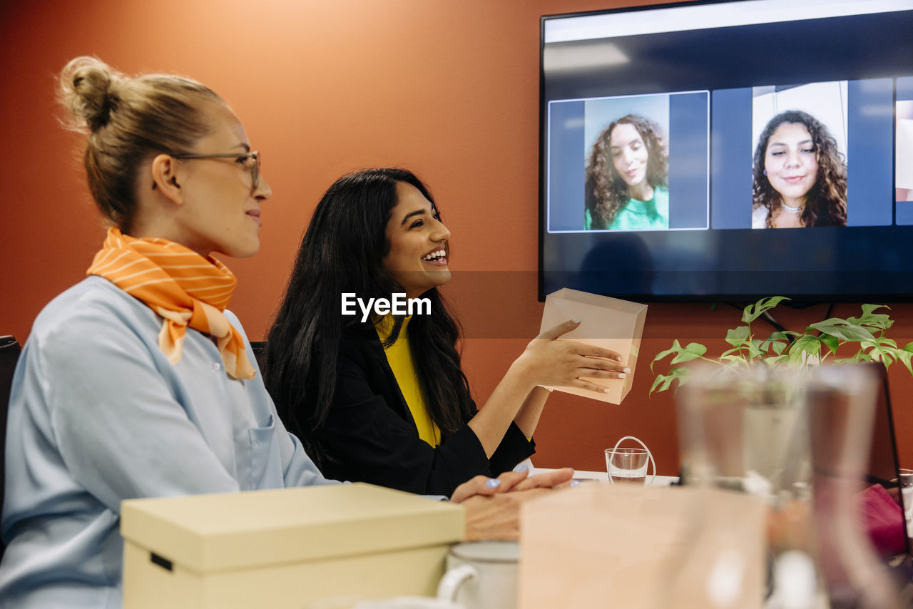 Happy businesswoman holding box during meeting with colleagues over video call at office