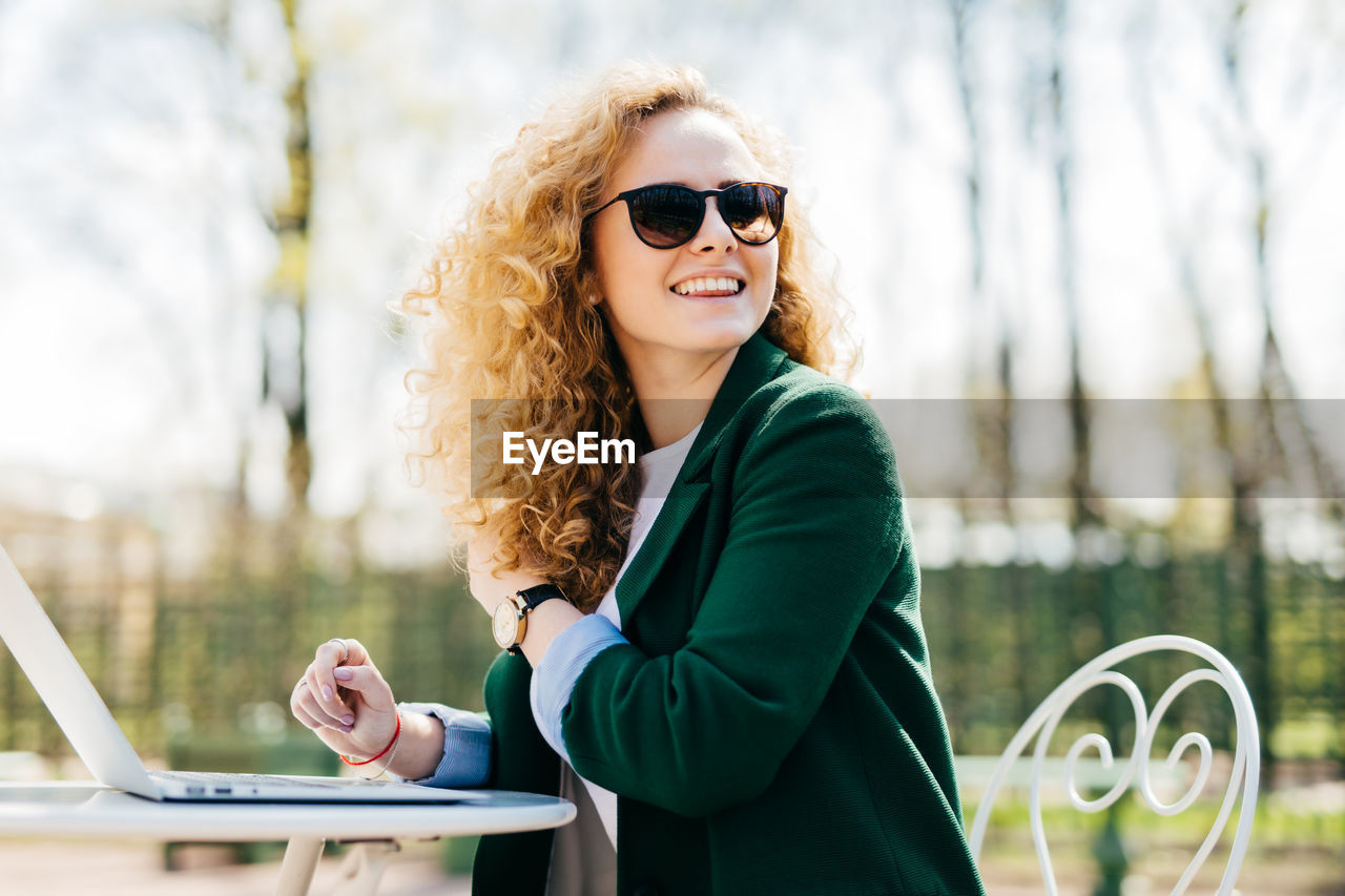 PORTRAIT OF SMILING YOUNG WOMAN WEARING SUNGLASSES