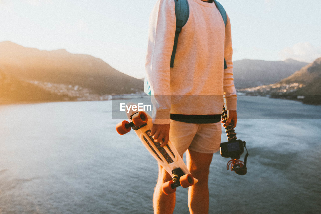 Midsection of man holding skateboard and camera against lake