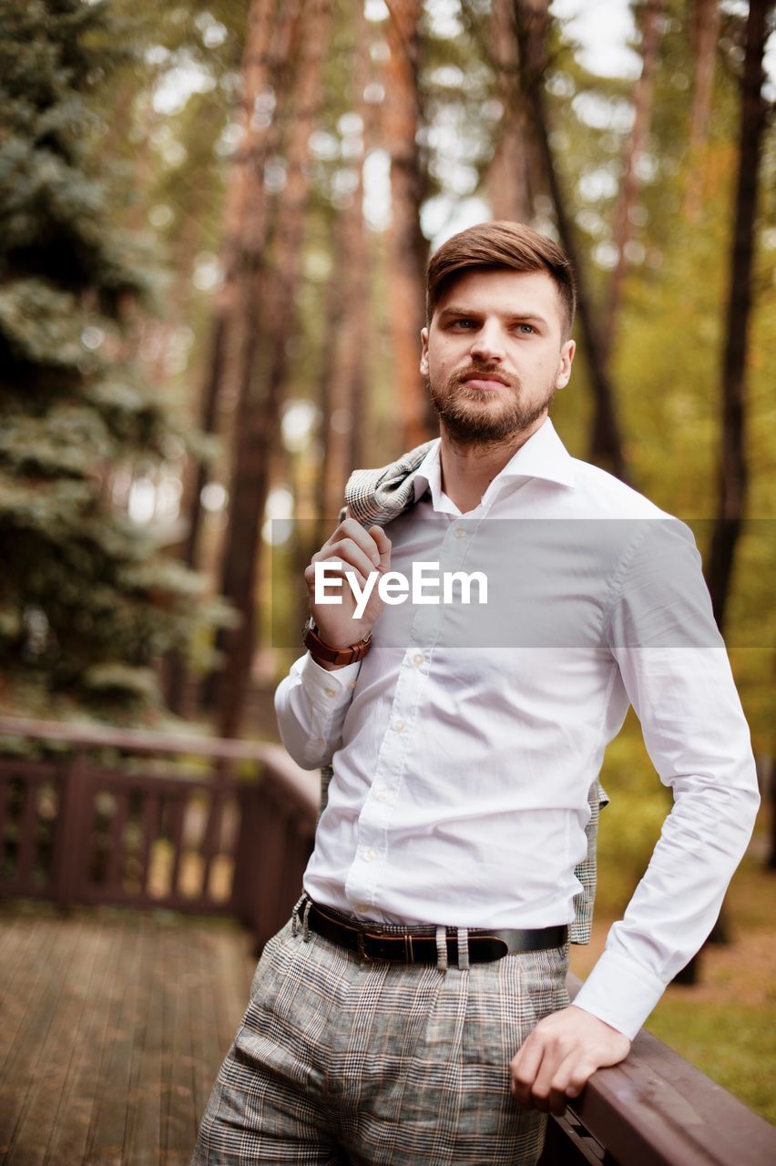 Young man looking away while standing by railing against trees in forest