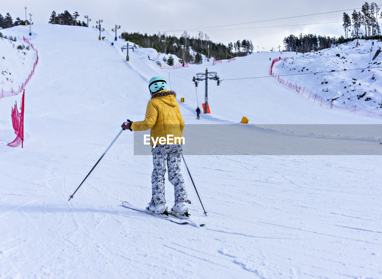 Rear view of young woman in yellow jacket and ski helmet skiing on mountain slope, winter sports