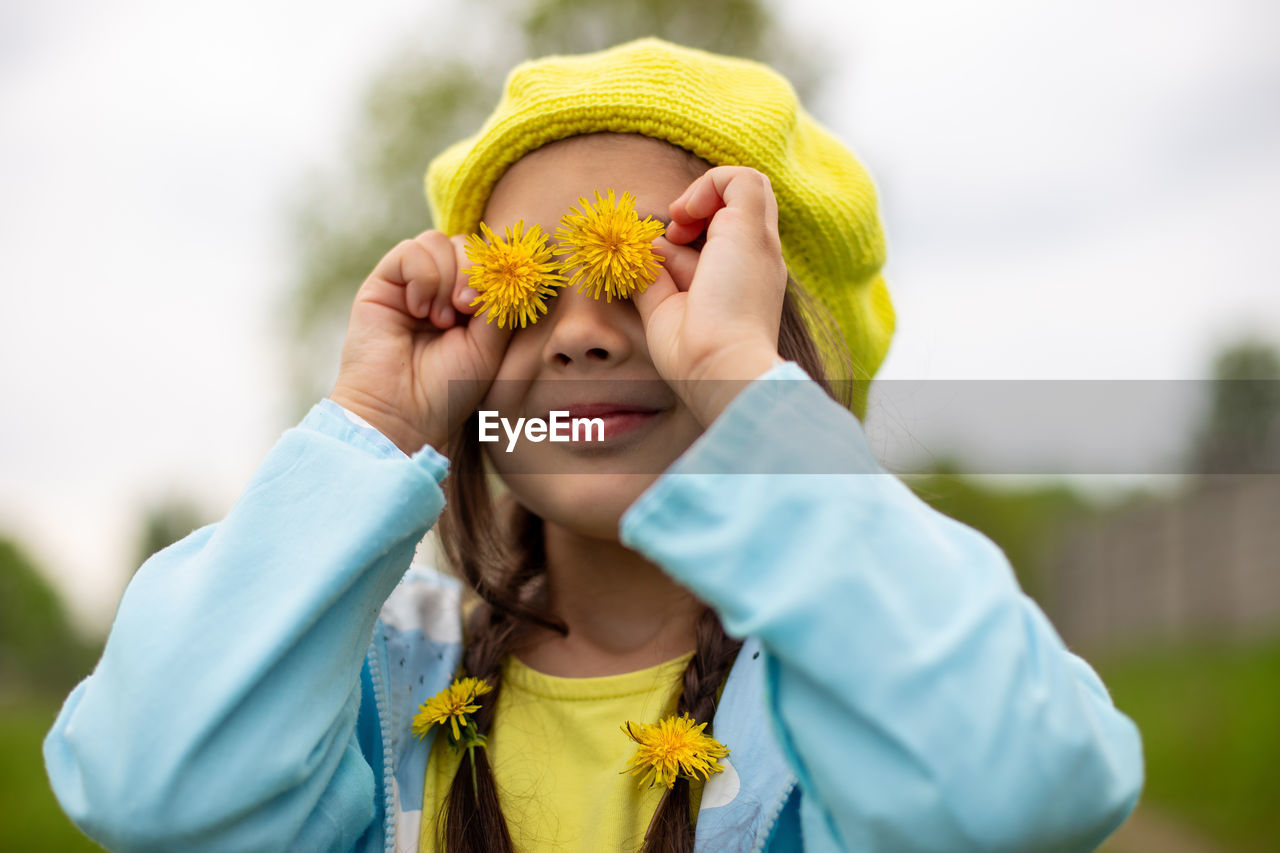 Portrait of a funny charming little girl with two yellow dandelion flowers near her eyes  in spring