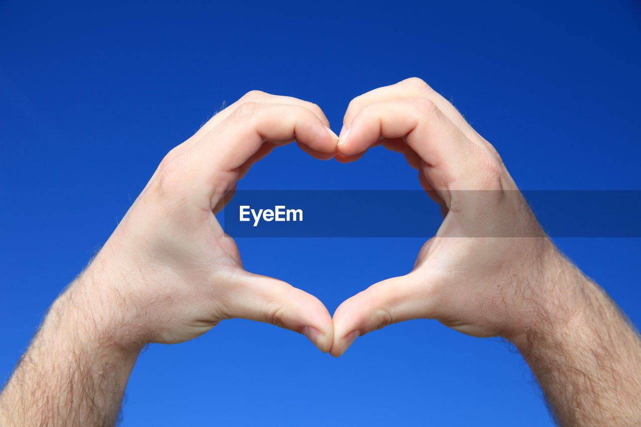 Cropped hand of man making heart shape against clear blue sky