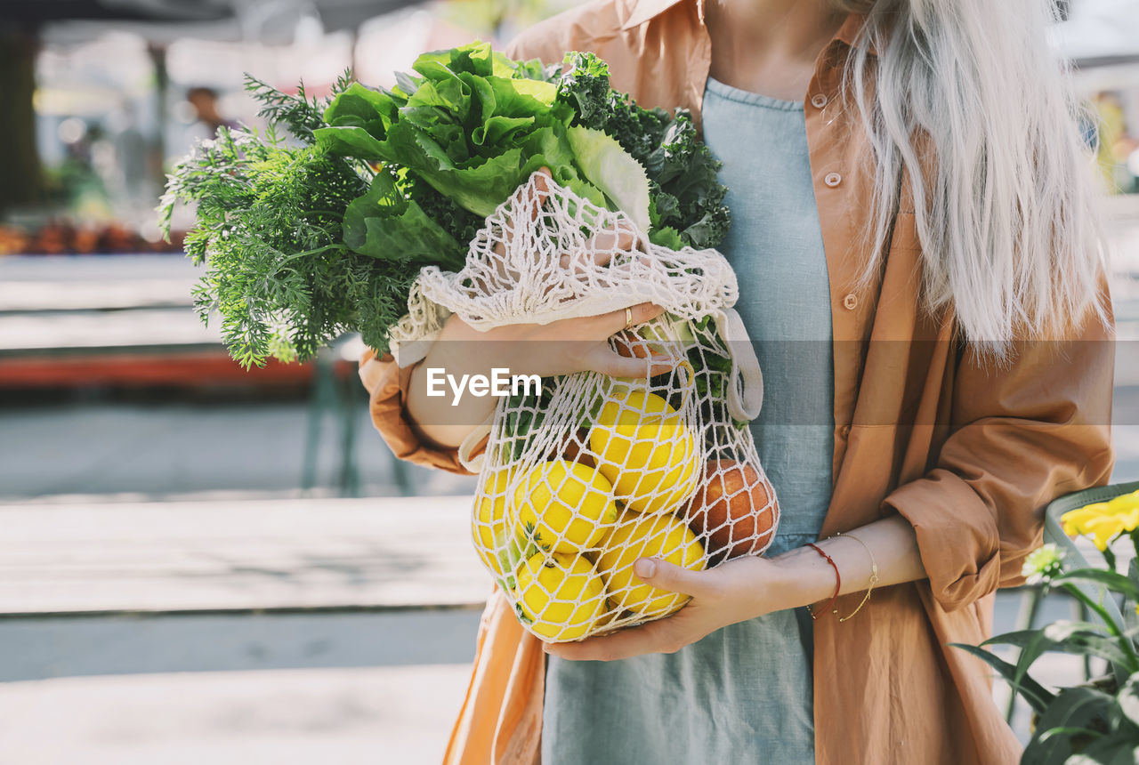 Customer holding leafy vegetables and lemons in mesh bag at local market