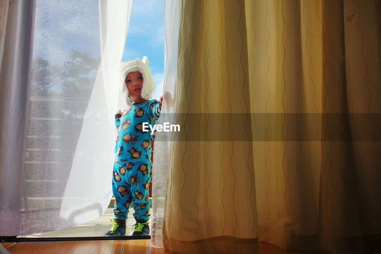 Full length of cute boy holding curtain while standing on balcony