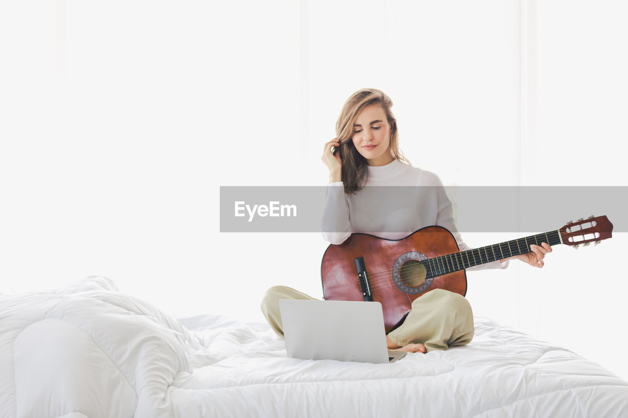 Full length of woman holding guitar by laptop while sitting on bed