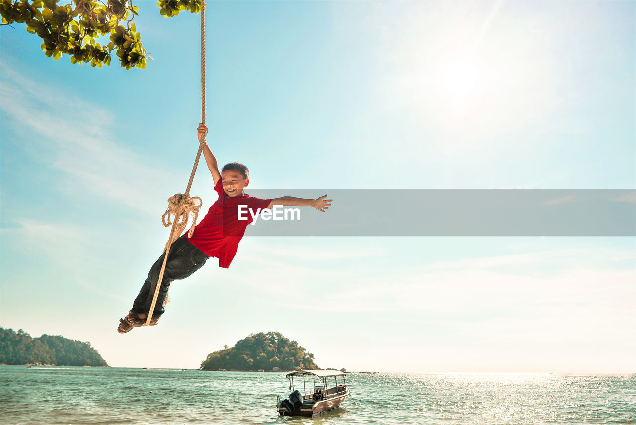 Boy swinging on rope swing over sea against sky during sunny day