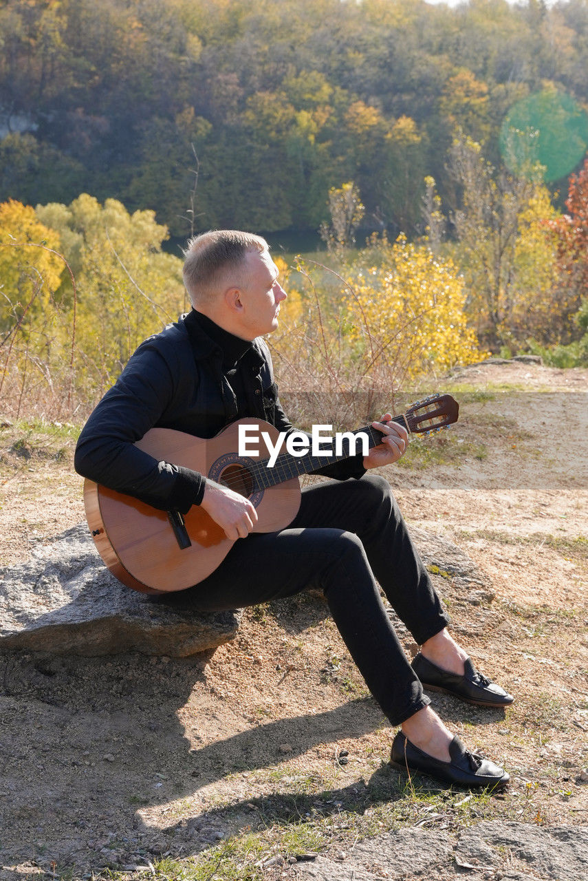 string instrument, musical instrument, one person, music, guitar, full length, musician, adult, sitting, leisure activity, arts culture and entertainment, plucking an instrument, nature, musical equipment, casual clothing, young adult, sunlight, day, lifestyles, acoustic guitar, person, men, tree, performance, plant, holding, outdoors, relaxation, skill, land, clothing, looking, concentration, side view