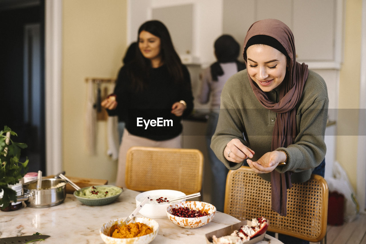 Smiling young woman in hijab garnishing food with pomegranate in kitchen at home