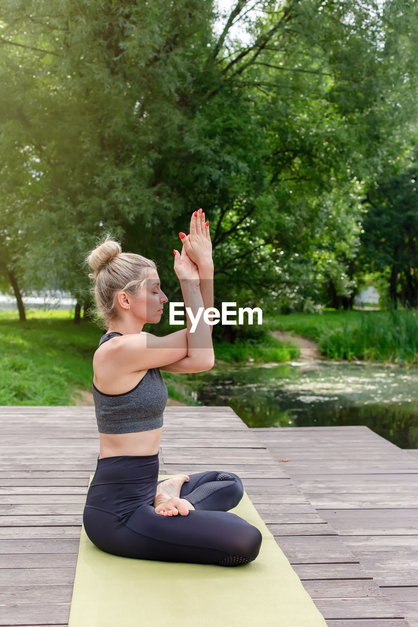 A beautiful woman sitting on a wooden platform by a pond on green mat in summer, does yoga