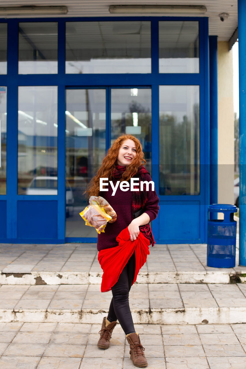 A cheerful red-haired young woman comes out of the store with bread