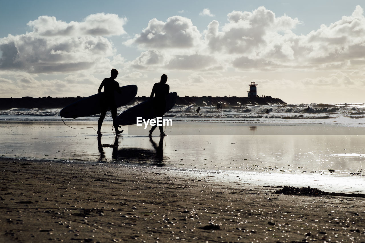 Silhouette surfers walking on shore at beach against sky