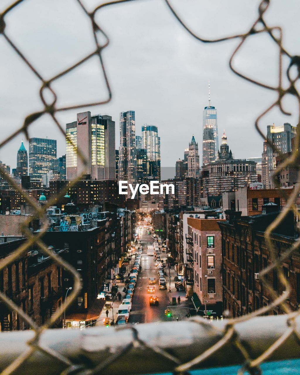 Road amidst buildings against cloudy sky seen through broken fence