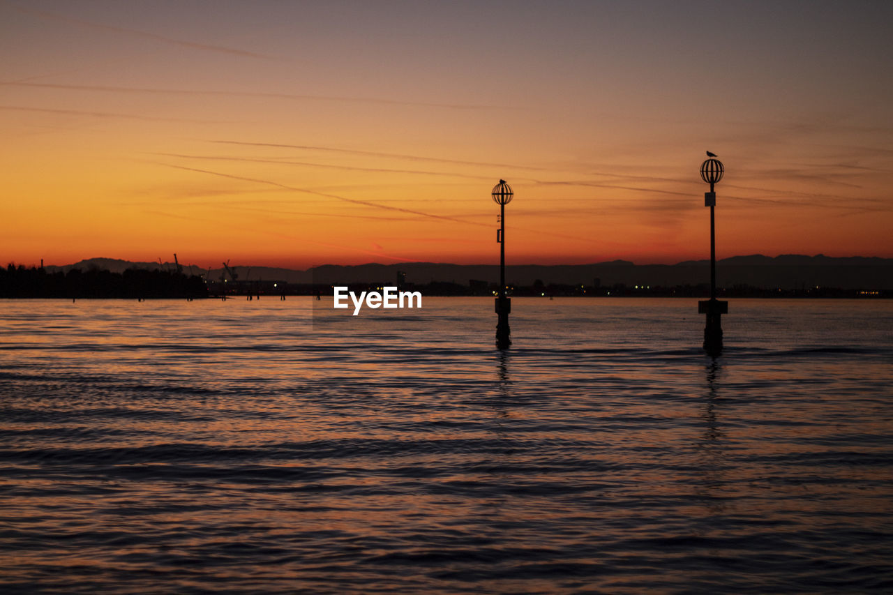 Silhouette street light by river against sky during sunset