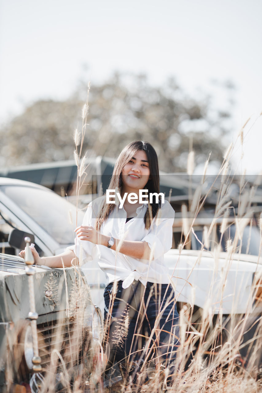 PORTRAIT OF YOUNG WOMAN STANDING ON CAR AGAINST SKY
