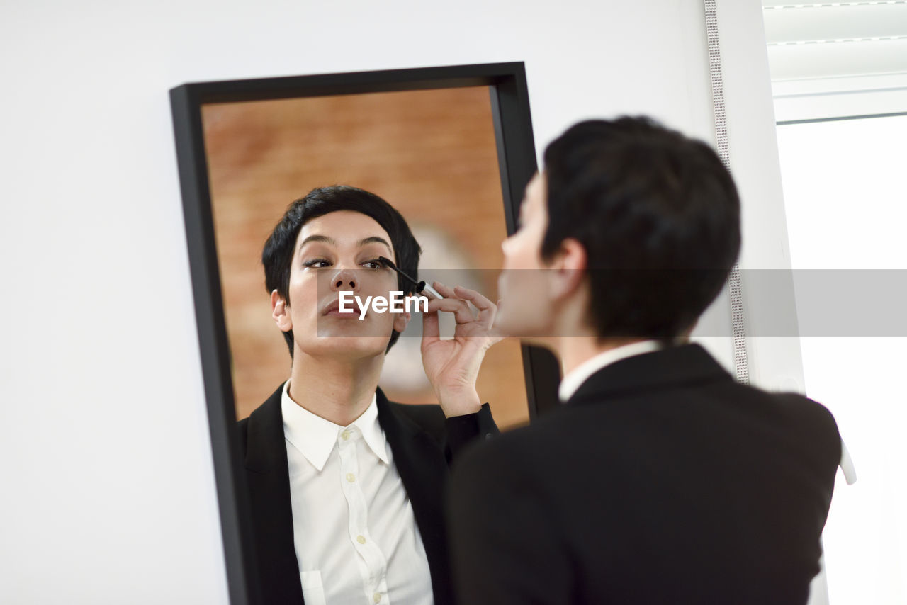 Woman applying mascara while standing against mirror