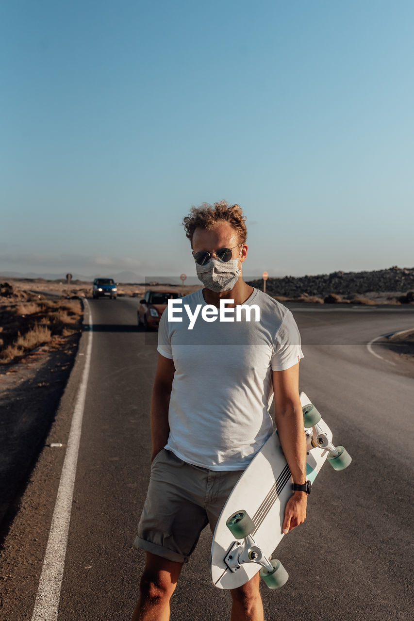 Portrait of man standing on road against clear sky with electric skateboard and mask