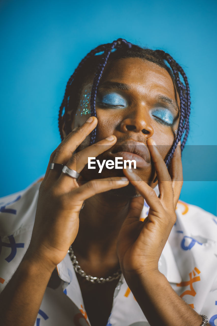 Serious african american male with braided hairstyle and bright makeup with closed eyes and hands on the face on blue background in studio