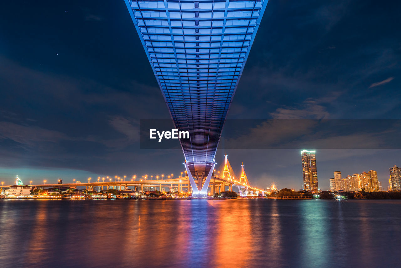 Illuminated bridge over river by buildings against sky at night