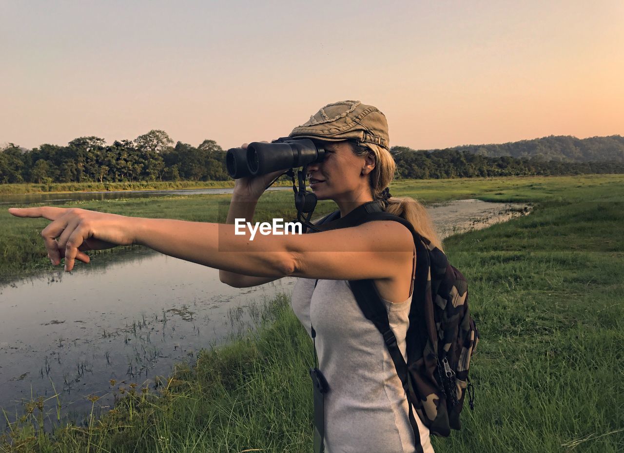 Woman standing on field while looking through binoculars against sky during sunset