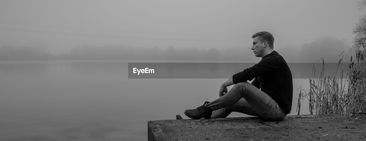 Young man sitting at lake against sky