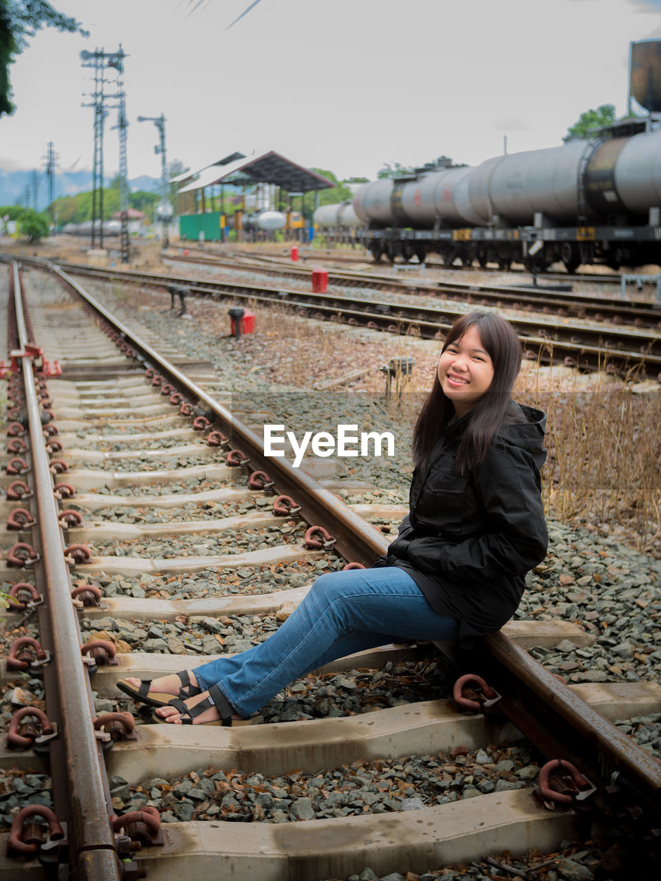 Portrait of smiling teenage girl sitting on railroad track against sky