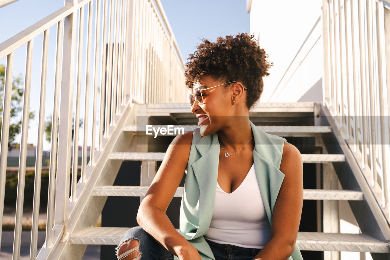 Low angle of cheerful young african american hipster female in trendy outfit and sunglasses sitting on stairs on urban street