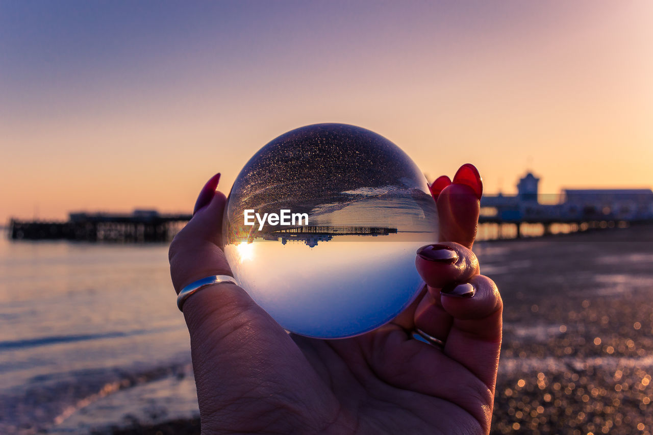 CLOSE-UP OF HAND HOLDING CRYSTAL BALL AGAINST SUNSET SKY
