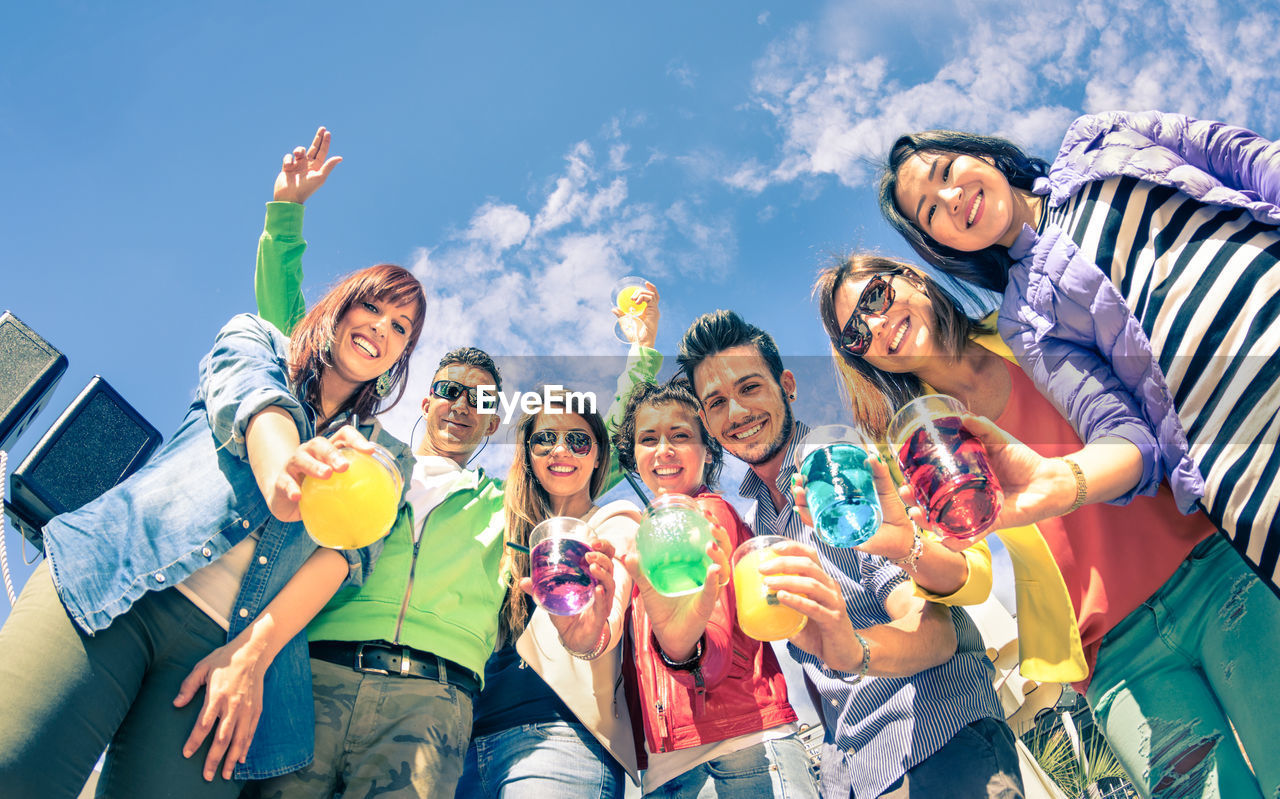 Low angle portrait of cheerful having drinks while standing against blue sky