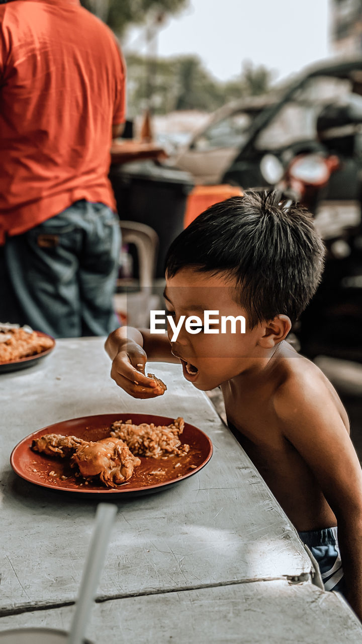 Side view of shirtless boy eating food while sitting on table