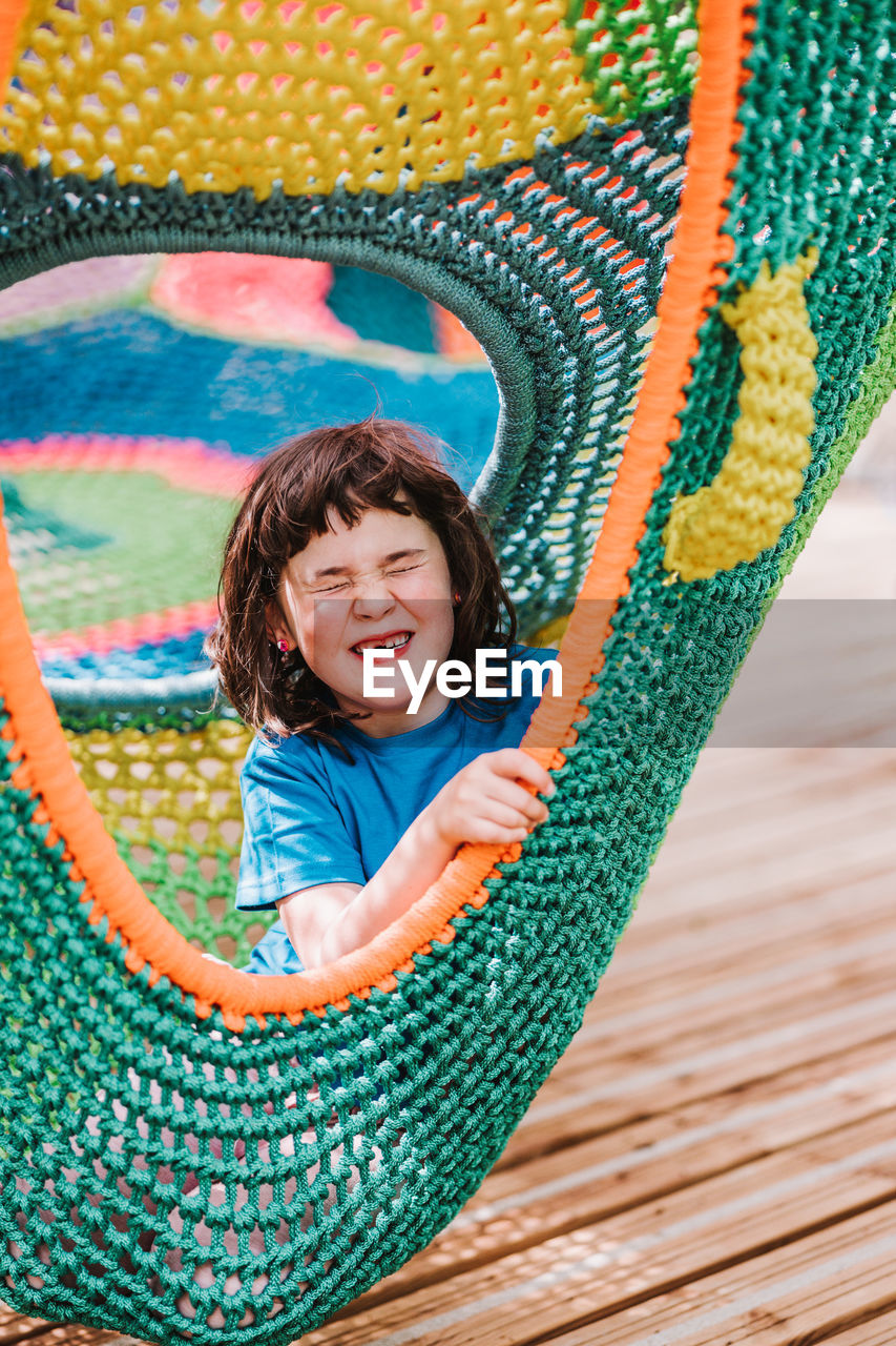 Adorable content little girl in t shirt smiling happily while relaxing with closed eyes inside of colorful knitted hammock in park