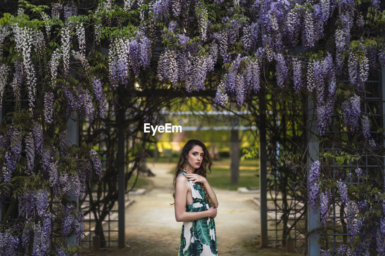 Side view attractive calm female in stylish sundress standing in verdant garden near blooming wistaria flowers while touching shoulder and looking at camera