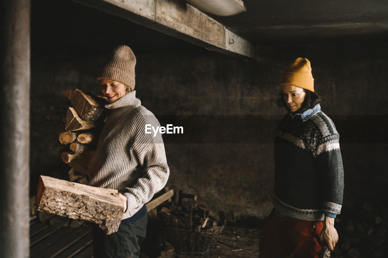 Mature woman looking at girlfriend collecting firewood while standing in shed