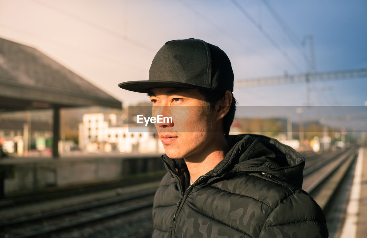 Thoughtful man wearing cap looking away at railroad station against sky