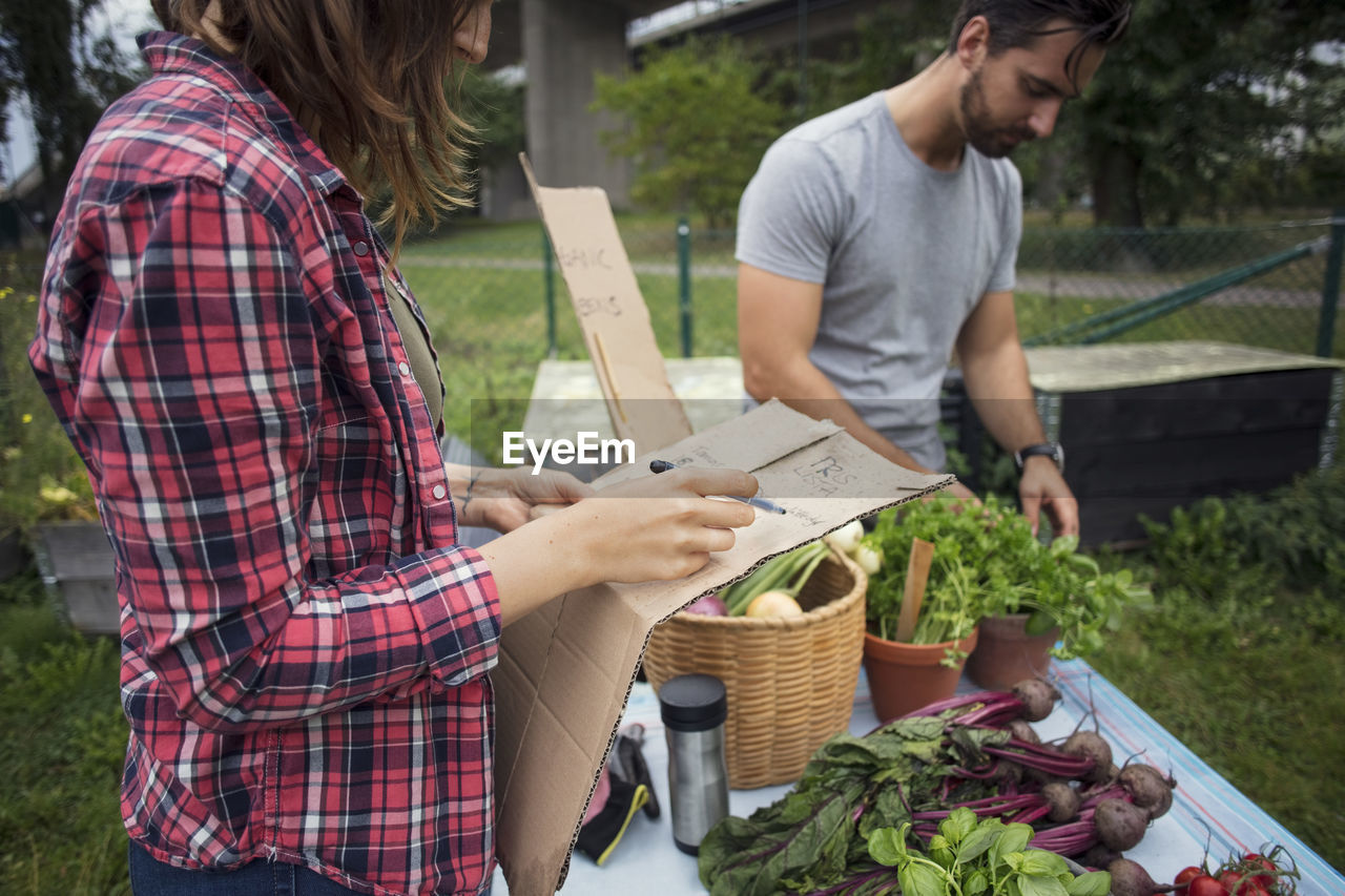 Midsection of woman writing on cardboard with man working in background at vegetable garden