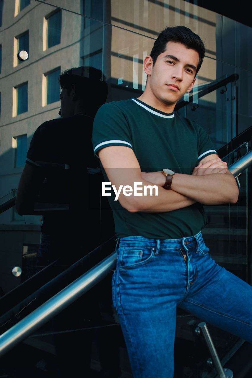 Portrait of young man with arms crossed standing against glass wall