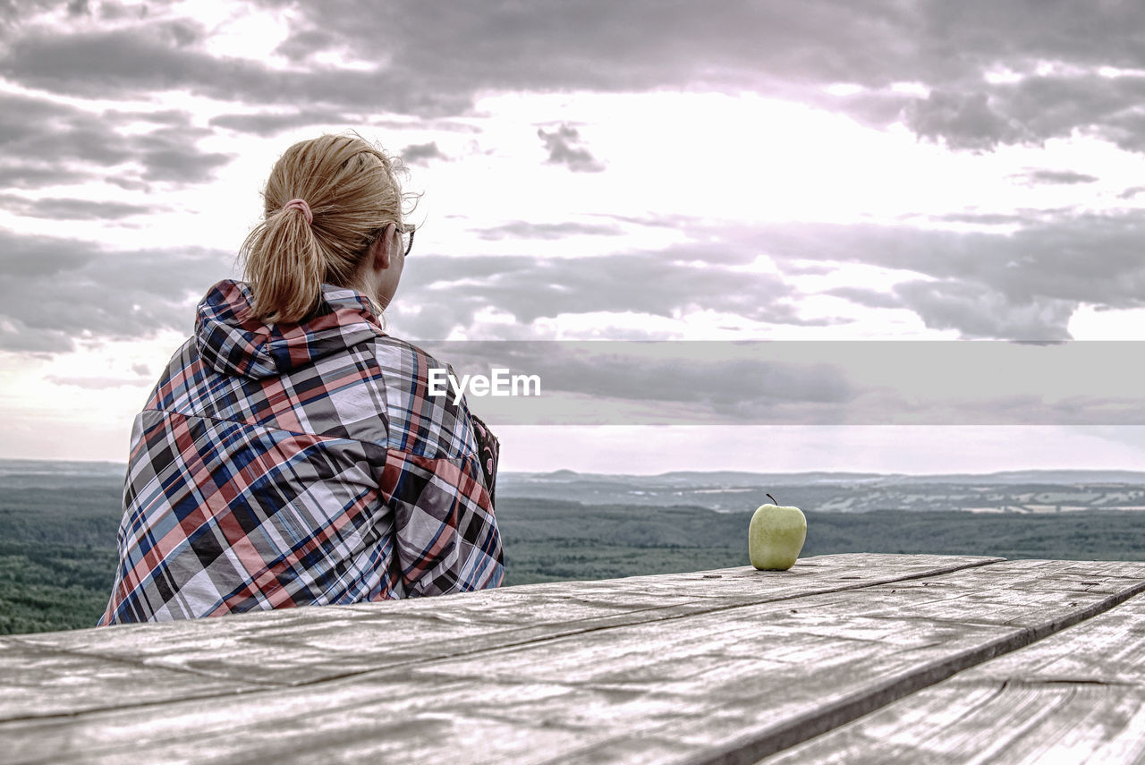 Young blond girl trekker resting at wooden table and taking break from hike. green apple on table