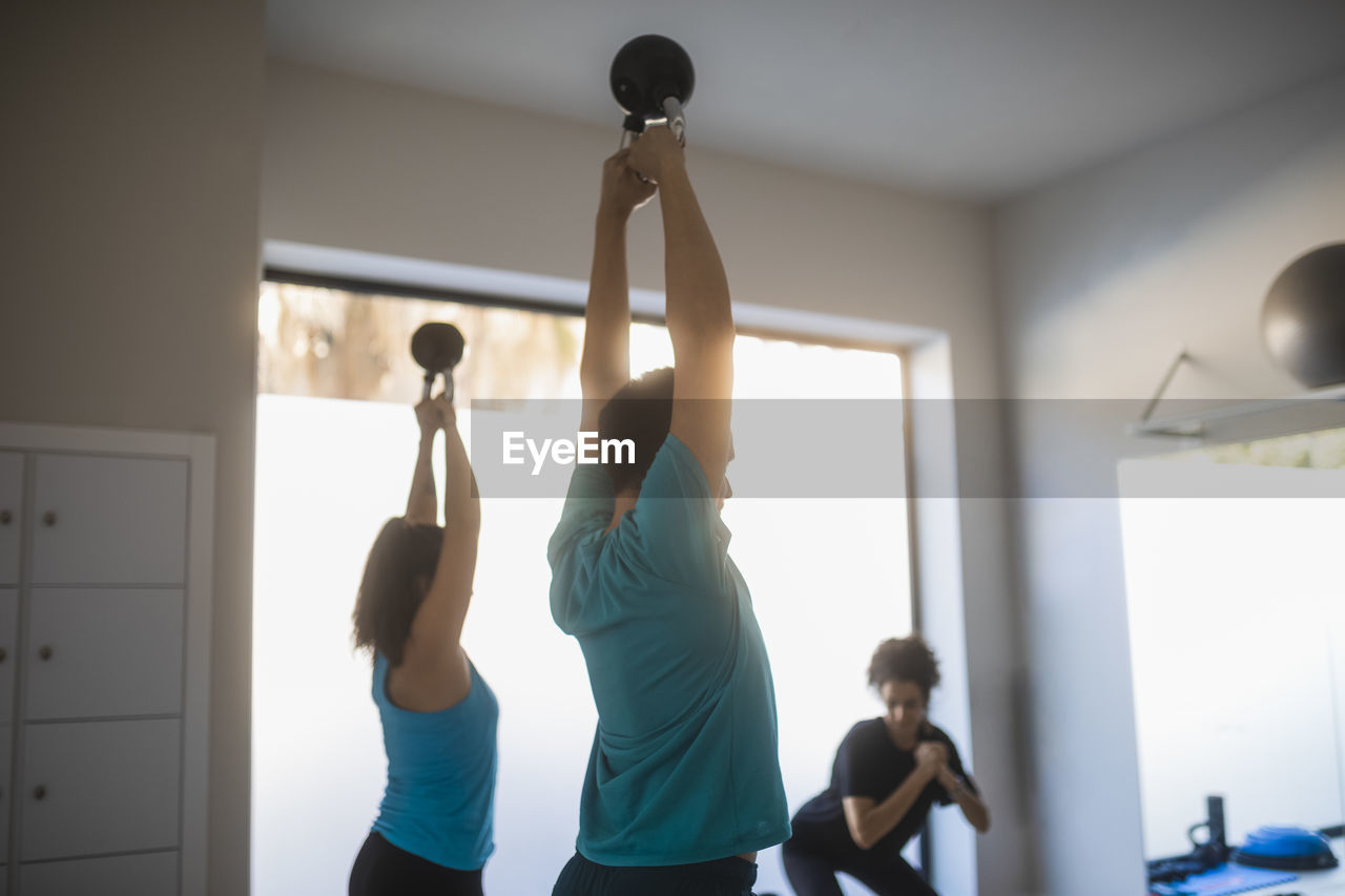 Side view of fit man and woman doing exercises with heavy kettlebells in raised hands during workout in fitness center