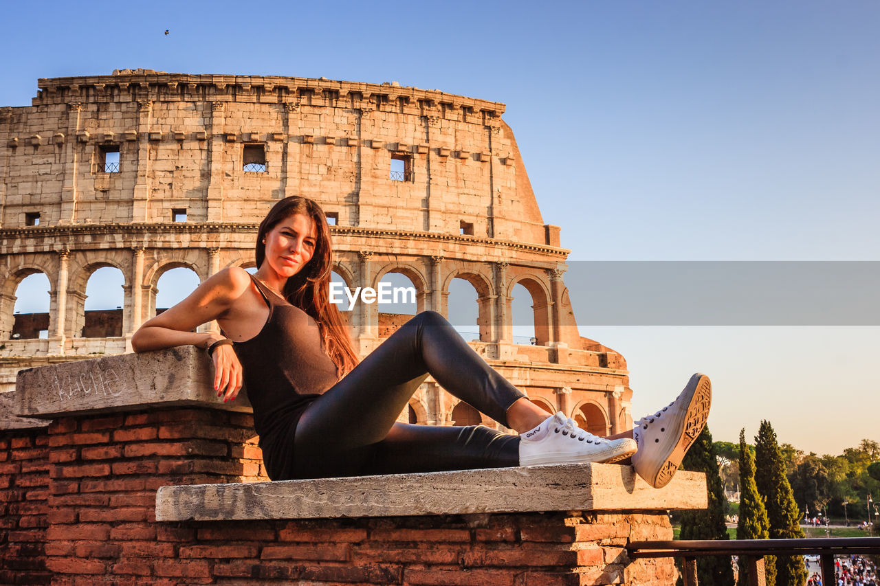 WOMAN SITTING ON HISTORICAL BUILDING AGAINST CLEAR SKY